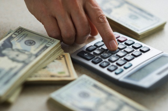 Close-up of man's hand using calculator and stacks of dollar bills --- Image by © Tetra Images/Corbis